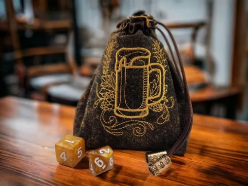 Dark brown fabric drawstring dice bag with gold-embroidered beer mug and hops design, next to two amber polyhedral dice on a wooden table.