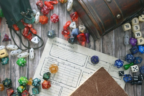 Overhead view of tabletop RPG equipment including a blank character sheet, a small wooden treasure chest, a green leather dice bag, and various colorful polyhedral dice.